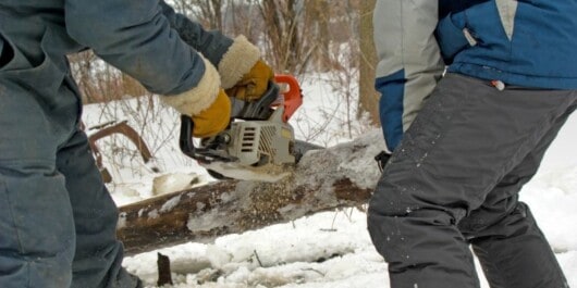 A person cutting log in winter using a chainsaw, and another person assisting in holding the log